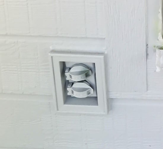 A close up of an outdoor electrical outlet on the side of a home with white siding. 