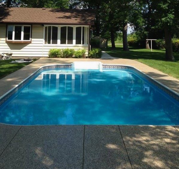 A backyard with a rectangular swimming pool with concrete surrounding. There is a house in the backg…