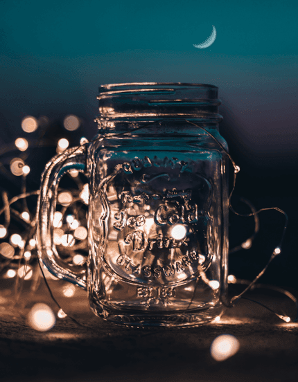 A mason jar with a handle in front of fairy lights laid out around the jar.