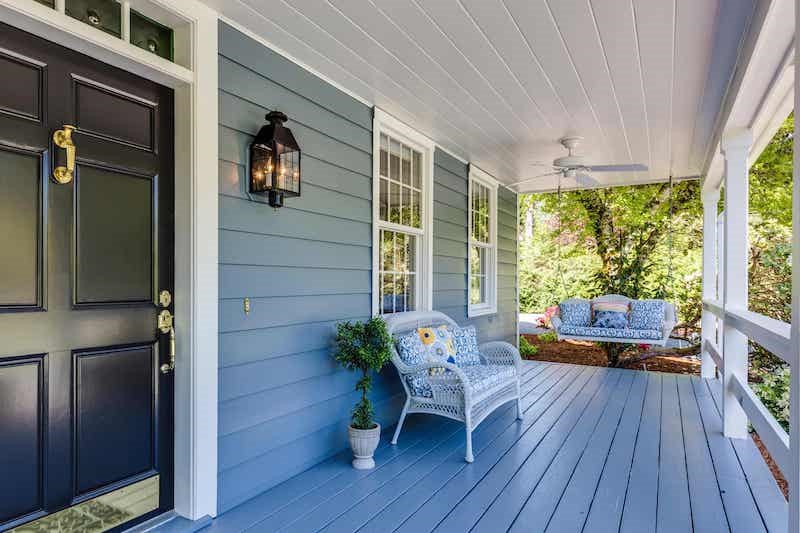 A front porch at an angle with a dark brown door on the left of the frame. The siding and porch are painted a light blue. There is a white whicker bench next to the door and a porch swing in the distance. There is lots of forage and greenery in the background.