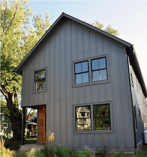 A house with vertical grey siding and wood posts in the front with black windows and a forest background.